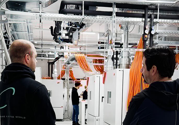 Personen stehen in einem Batterietestlabor mit BINDER Batterietestschränken. An der Decke verlaufen zahlreiche orangefarbene Kabelstränge, silberne Lüftungsrohre und Leitungen. Zwei Personen im Vordergrund betrachten die Anlage, während weitere Personen im Hintergrund an den Geräten arbeiten.