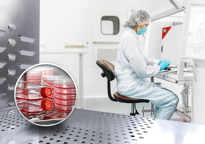 View into a CO2 incubator with cell culture bottles. In the background, a laboratory employee in protective clothing working at a safety cabinet.