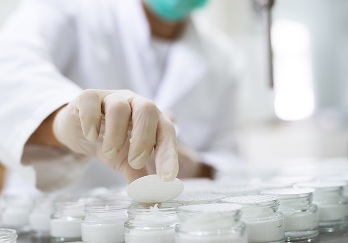 Cosmetic cream samples in a lab. Lab worker wearing gloves and face mask in the background.